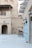 A traditional Middle Eastern street scene with sand-colored buildings and wooden beams jutting from the walls. Small balconies with latticework are adorned with flags. The courtyard-like setting is lined with blue doors on one side and features a wooden cart with metal pots in the foreground. The area is lit by several wall-mounted lanterns.