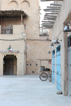 A traditional Middle Eastern street scene with sand-colored buildings and wooden beams jutting from the walls. Small balconies with latticework are adorned with flags. The courtyard-like setting is lined with blue doors on one side and features a wooden cart with metal pots in the foreground. The area is lit by several wall-mounted lanterns.