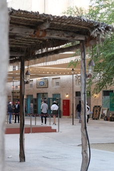 A quaint outdoor setting featuring a rustic wooden canopy with a thatched roof. Several people stand in the background near an art gallery, depicted by paintings leaning against the wall, and a sign in Arabic. The courtyard area has string lights hanging above, adding to the decorative atmosphere.