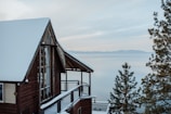 A rustic wooden cabin with a snow-covered roof is positioned near a calm, expansive body of water. In the background, distant mountains are visible under a soft, overcast sky. Pine trees, also dusted with snow, frame the scene, creating a tranquil and serene atmosphere.