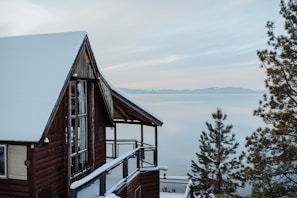 A rustic wooden cabin with a snow-covered roof is positioned near a calm, expansive body of water. In the background, distant mountains are visible under a soft, overcast sky. Pine trees, also dusted with snow, frame the scene, creating a tranquil and serene atmosphere.