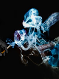Intricate swirls of blue and white smoke rise and twist against a dark background, originating from a lit cigarette held in a hand.