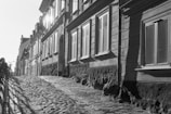 Black and white photograph of an old cobblestone street wet from rain, lined with austere historic buildings.