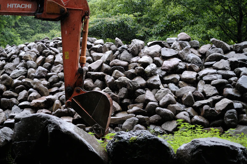 Excavator setting a basalt boulder into a terraced retaining wall on a sloped Bend lot