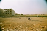 Several children are playing on a large, open sandy field with a few small patches of grass. In the background, there are unfinished or abandoned buildings, and the atmosphere seems somewhat desolate. The children are in action, running and scattered across the foreground, suggesting a game or playful activity.