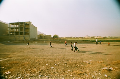 Several children are playing on a large, open sandy field with a few small patches of grass. In the background, there are unfinished or abandoned buildings, and the atmosphere seems somewhat desolate. The children are in action, running and scattered across the foreground, suggesting a game or playful activity.