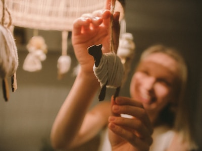 A person is happily holding a small, handcrafted woolen sheep hanging from a fixture. The background is softly lit and out of focus, with other similar woolen objects visible.