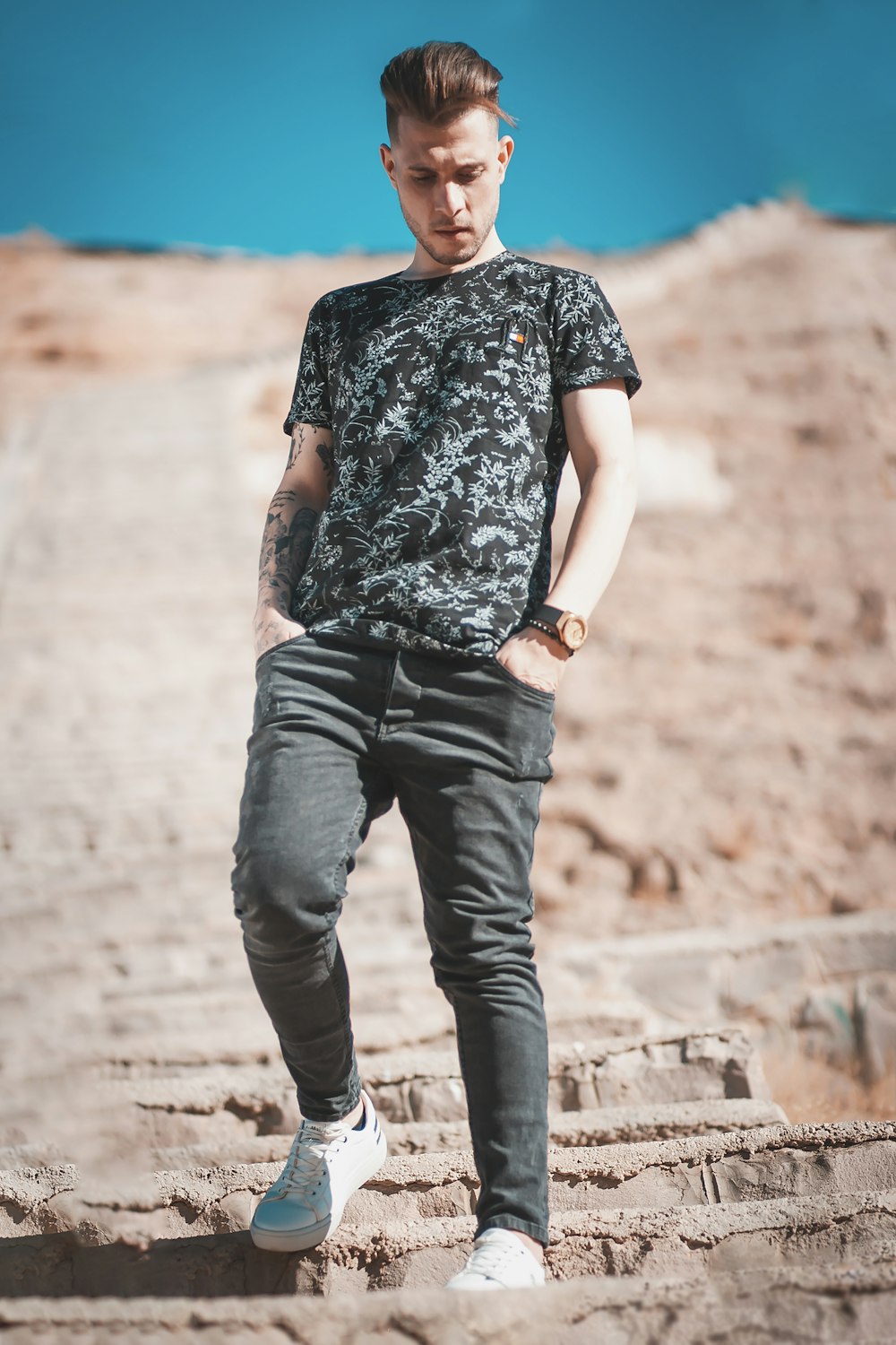 man in black and white floral shirt and blue denim jeans standing on brown concrete pavement