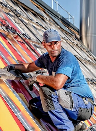 A worker in a blue shirt and cap is installing a solar panel on a slanted rooftop with bright colored panels. He is wearing gloves and appears focused on the task, with various tools and wires visible around him.