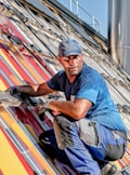 A worker in a blue shirt and cap is installing a solar panel on a slanted rooftop with bright colored panels. He is wearing gloves and appears focused on the task, with various tools and wires visible around him.