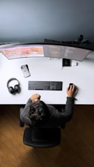 A person is sitting at a desk with a dual curved monitor setup. The desk is organized with a keyboard, mouse, smartphone, and a pair of headphones. The person seems to be working on coding projects as lines of code are visible on the screens.