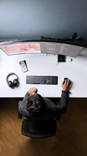 A person is sitting at a desk with a dual curved monitor setup. The desk is organized with a keyboard, mouse, smartphone, and a pair of headphones. The person seems to be working on coding projects as lines of code are visible on the screens.