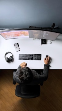 A person is sitting at a desk with a dual curved monitor setup. The desk is organized with a keyboard, mouse, smartphone, and a pair of headphones. The person seems to be working on coding projects as lines of code are visible on the screens.