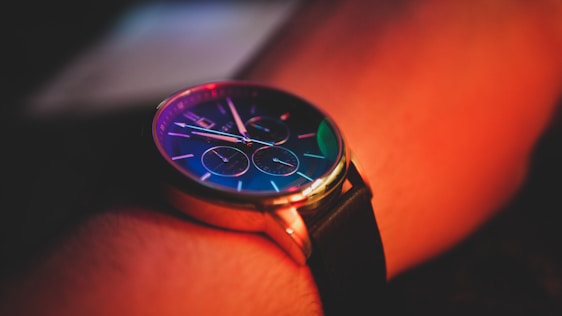 A wristwatch with a round face and a black leather strap is visible, reflecting vibrant colors. The watch face displays several dials and is illuminated by a red and orange ambient light, creating a warm glow on the wrist.