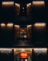 The image depicts a traditional building at night, featuring muted warm lighting highlighting intricately designed facade elements. There is a red illuminated sign with characters above an arch entryway. The upper levels showcase lattice windows with ambient light, adding a moody and atmospheric quality.