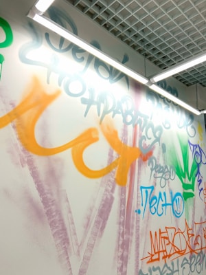 A wall with colorful graffiti featuring various abstract shapes and text in different languages, predominantly in shades of orange, purple, green, and blue. The graffiti is illuminated by overhead fluorescent lights and surrounded by a modern ceiling grid design.
