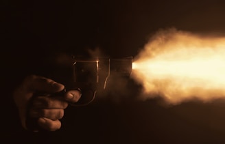 A hand is holding a revolver, which is firing. A bright muzzle flash is visible, illuminating the otherwise dark scene.
