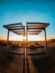 Two wooden pergolas stand on a deck with long shadows extending toward the viewer. The sun sets between the structures, casting warm golden light over the boardwalk and surrounding grassy area. A clear blue sky gradually deepens in color towards the top of the image.