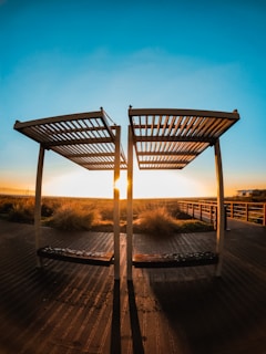 Two wooden pergolas stand on a deck with long shadows extending toward the viewer. The sun sets between the structures, casting warm golden light over the boardwalk and surrounding grassy area. A clear blue sky gradually deepens in color towards the top of the image.