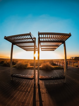 Two wooden pergolas stand on a deck with long shadows extending toward the viewer. The sun sets between the structures, casting warm golden light over the boardwalk and surrounding grassy area. A clear blue sky gradually deepens in color towards the top of the image.