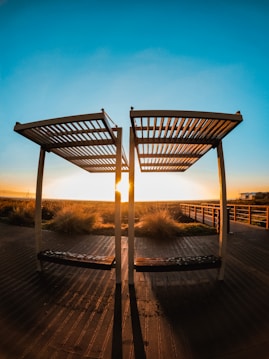 Two wooden pergolas stand on a deck with long shadows extending toward the viewer. The sun sets between the structures, casting warm golden light over the boardwalk and surrounding grassy area. A clear blue sky gradually deepens in color towards the top of the image.