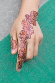 A hand adorned with intricate henna designs featuring floral and paisley patterns. The artwork extends from the forefinger up across the back of the hand. The henna is rich in color and detailed with dot accents.