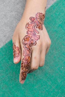 A hand adorned with intricate henna designs featuring floral and paisley patterns. The artwork extends from the forefinger up across the back of the hand. The henna is rich in color and detailed with dot accents.