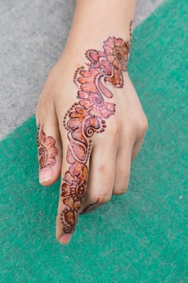 A hand adorned with intricate henna designs featuring floral and paisley patterns. The artwork extends from the forefinger up across the back of the hand. The henna is rich in color and detailed with dot accents.