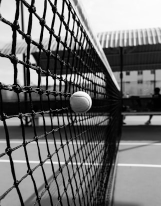 A tennis ball is captured mid-air as it is stopped by a tennis net. The background shows a partially blurred tennis court with some indistinguishable structures and possibly a person or player.