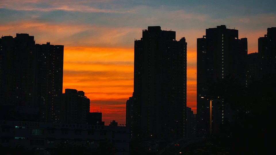 The image depicts a city skyline at sunset, with tall buildings silhouetted against an orange sky. The sun is setting behind the buildings, casting long shadows and creating a warm glow over the scene. The perspective is from below, looking up towards the towering structures that dominate the frame.