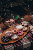 A rustic holiday-themed table setting featuring an assortment of colorful decorated donuts on a wooden platter. The table is covered with a green, red, and white plaid cloth. In the background, there are small, decorated gingerbread houses and potted mini Christmas trees wrapped with burlap.