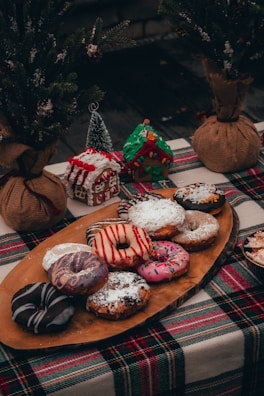A rustic holiday-themed table setting featuring an assortment of colorful decorated donuts on a wooden platter. The table is covered with a green, red, and white plaid cloth. In the background, there are small, decorated gingerbread houses and potted mini Christmas trees wrapped with burlap.