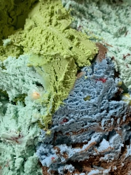 A close-up view of multiple scoops of ice cream in various colors, including green, blue, and brown. The ice cream appears textured and creamy, with bits of different colors embedded throughout.