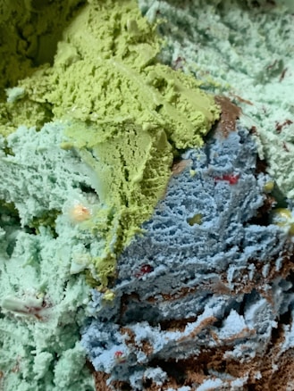 A close-up view of multiple scoops of ice cream in various colors, including green, blue, and brown. The ice cream appears textured and creamy, with bits of different colors embedded throughout.