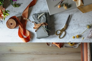 A beautifully wrapped gift lies on a marble surface, adorned with dried flowers and tied with a brown ribbon. Nearby, a pair of vintage-style gold scissors, a roll of orange ribbon, and additional decorative floral elements are arranged in an artistic manner. The table has a clean, elegant look with scattered natural accents, and the lower section of the image shows a wooden floor.