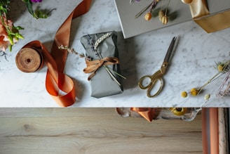 A beautifully wrapped gift lies on a marble surface, adorned with dried flowers and tied with a brown ribbon. Nearby, a pair of vintage-style gold scissors, a roll of orange ribbon, and additional decorative floral elements are arranged in an artistic manner. The table has a clean, elegant look with scattered natural accents, and the lower section of the image shows a wooden floor.