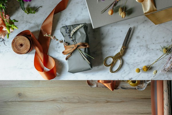 A beautifully wrapped gift lies on a marble surface, adorned with dried flowers and tied with a brown ribbon. Nearby, a pair of vintage-style gold scissors, a roll of orange ribbon, and additional decorative floral elements are arranged in an artistic manner. The table has a clean, elegant look with scattered natural accents, and the lower section of the image shows a wooden floor.