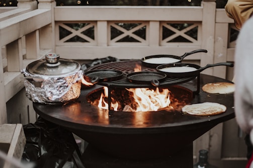 A large outdoor cast iron fire pit with an open flame is being used for cooking. Multiple cast iron pans are positioned on a grill above the fire, with one covered pot and a couple of pancakes either cooking or resting on the sides. The scene is enclosed by a light-colored wooden fence.