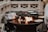 A large outdoor cast iron fire pit with an open flame is being used for cooking. Multiple cast iron pans are positioned on a grill above the fire, with one covered pot and a couple of pancakes either cooking or resting on the sides. The scene is enclosed by a light-colored wooden fence.