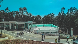 A modern building with a green roof surrounded by tall trees. People are walking along a slightly curved road next to the building, some holding umbrellas. The sky is a clear blue, and there is signage near the building entrance. The area appears to be landscaped with pavement and some decorative elements.