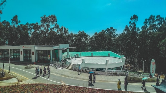 A modern building with a green roof surrounded by tall trees. People are walking along a slightly curved road next to the building, some holding umbrellas. The sky is a clear blue, and there is signage near the building entrance. The area appears to be landscaped with pavement and some decorative elements.