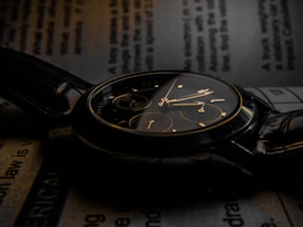 A close-up view of an elegant black wristwatch with gold accents resting on an open book. The watch face includes multiple dials and is slightly illuminated, creating a sophisticated and classic aesthetic. The surrounding text on the book is blurred, drawing attention to the timepiece.