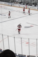 Several hockey players in red and white uniforms are positioned on an ice rink, preparing for a play. The scene is viewed through a net, adding a grid-like filter to the image. Advertisements and audience members are visible on the far side of the rink.