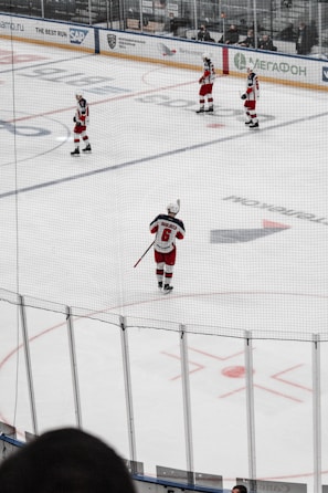 Several hockey players in red and white uniforms are positioned on an ice rink, preparing for a play. The scene is viewed through a net, adding a grid-like filter to the image. Advertisements and audience members are visible on the far side of the rink.