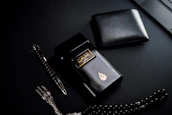 A dark-themed flat lay featuring a black perfume bottle with gold Arabic script and detailing, placed beside a black wallet. A sleek black and silver pen and a string of black prayer beads with a silver tassel complement the arrangement. The overall setup is positioned against a matte black background.