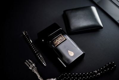 A dark-themed flat lay featuring a black perfume bottle with gold Arabic script and detailing, placed beside a black wallet. A sleek black and silver pen and a string of black prayer beads with a silver tassel complement the arrangement. The overall setup is positioned against a matte black background.
