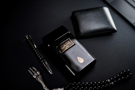 A dark-themed flat lay featuring a black perfume bottle with gold Arabic script and detailing, placed beside a black wallet. A sleek black and silver pen and a string of black prayer beads with a silver tassel complement the arrangement. The overall setup is positioned against a matte black background.