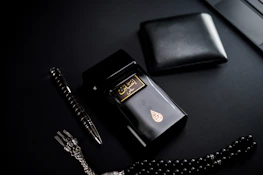 A dark-themed flat lay featuring a black perfume bottle with gold Arabic script and detailing, placed beside a black wallet. A sleek black and silver pen and a string of black prayer beads with a silver tassel complement the arrangement. The overall setup is positioned against a matte black background.