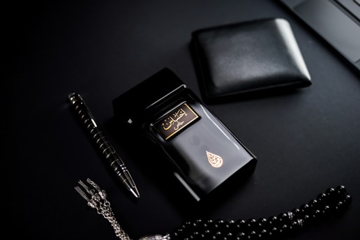 A dark-themed flat lay featuring a black perfume bottle with gold Arabic script and detailing, placed beside a black wallet. A sleek black and silver pen and a string of black prayer beads with a silver tassel complement the arrangement. The overall setup is positioned against a matte black background.