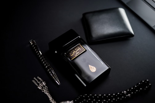 A dark-themed flat lay featuring a black perfume bottle with gold Arabic script and detailing, placed beside a black wallet. A sleek black and silver pen and a string of black prayer beads with a silver tassel complement the arrangement. The overall setup is positioned against a matte black background.